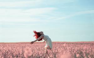 A vibrant woman with fiery red hair, dancing in a field of fluffy dandelion-like flowers. She wears a flowing white sun dress, her body arched backward, arms gracefully stretched behind her, against a serene blue sky with wispy clouds