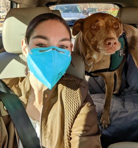Woman buckled into seat of car with a mask on, with service dog buckled in the back seat.