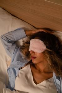 Woman sleeping with one arm over head with pink fuzzy eye mask in blue long sleeve shirt and white under shirt.