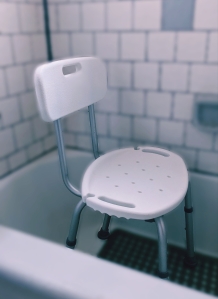 White shower chair sitting in a white tub with dark grey bath matt and white tile and dark grey grout.