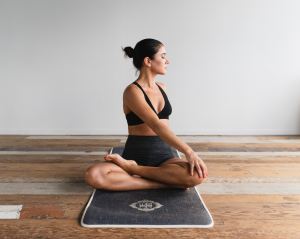 Woman sitting on hardwood floor, legs crossed, with her body gently rotated as she stretches her back. One hand is resting on her left knee, and her eyes are closed, looking relaxed. Yoga mat beneath her, with a light grey wall in the background.