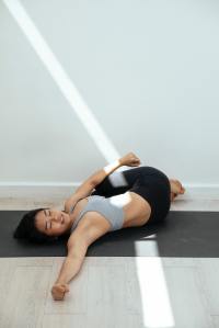 Fit woman lying on a yoga mat, performing a supine spinal twist with a serene smile on her face. A soft beam of light from an out-of-frame window illuminates the wall and gently crosses her body, adding a dynamic touch to the tranquil scene